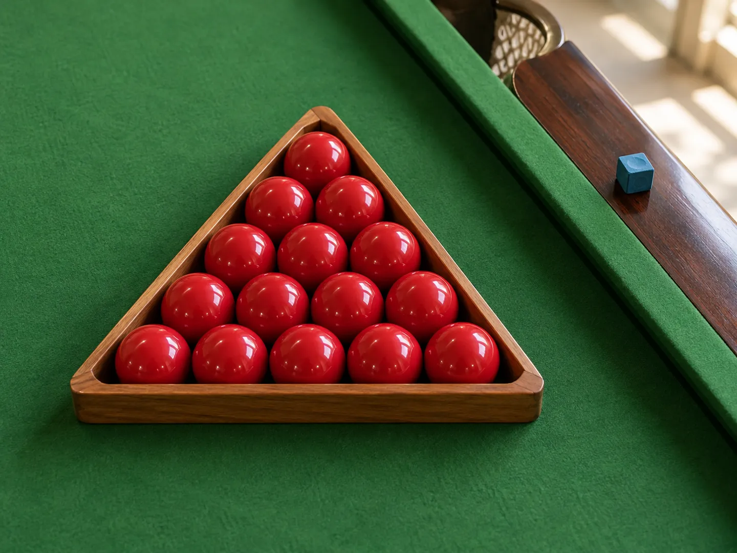 Fifteen red snooker balls packed in a wooden triangle on green cloth, with a small blue chalk cube on the polished wooden rail