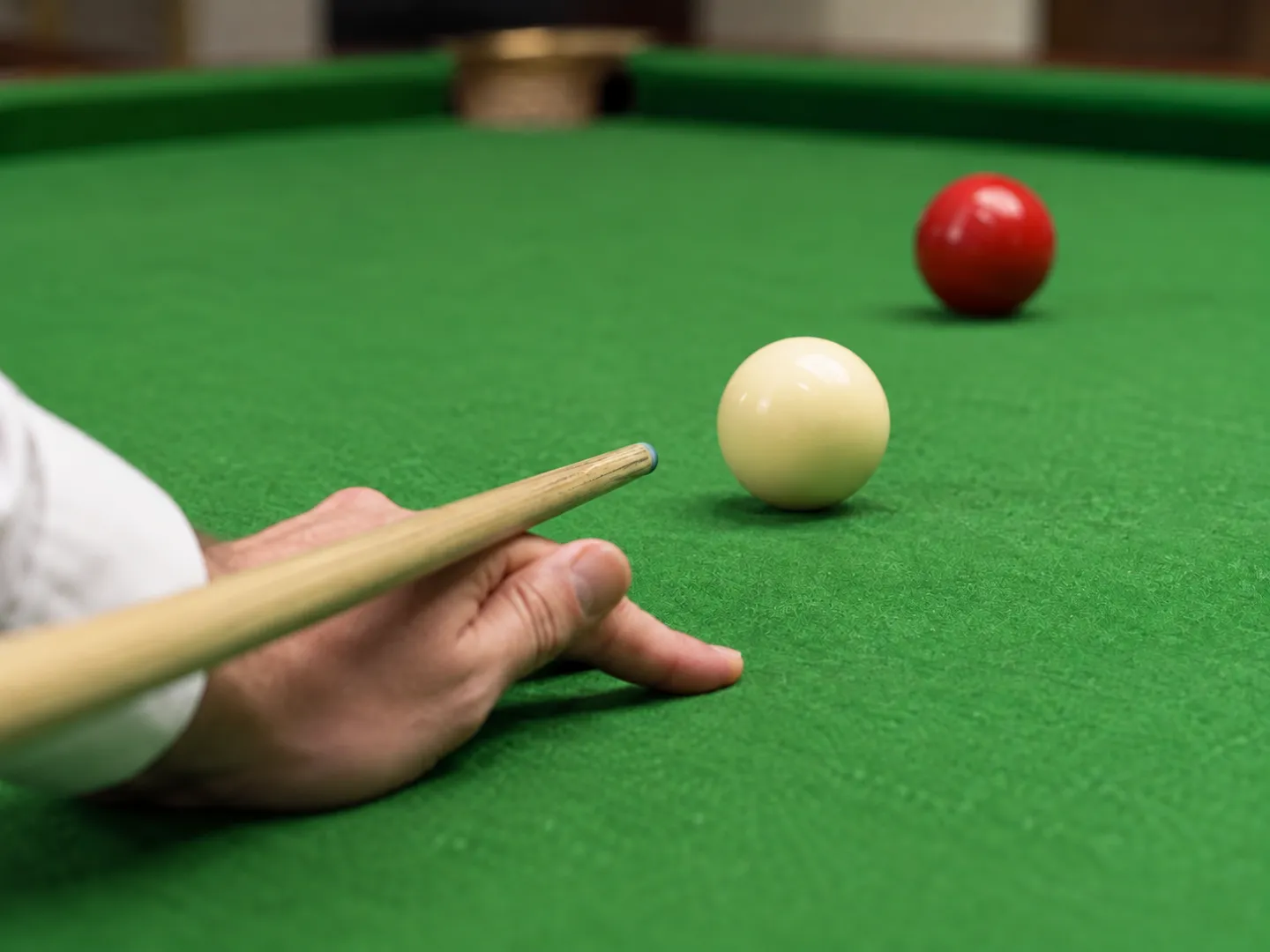 A hand forming a bridge on green cloth with a wooden cue lined up behind a white cue ball and a red ball beyond